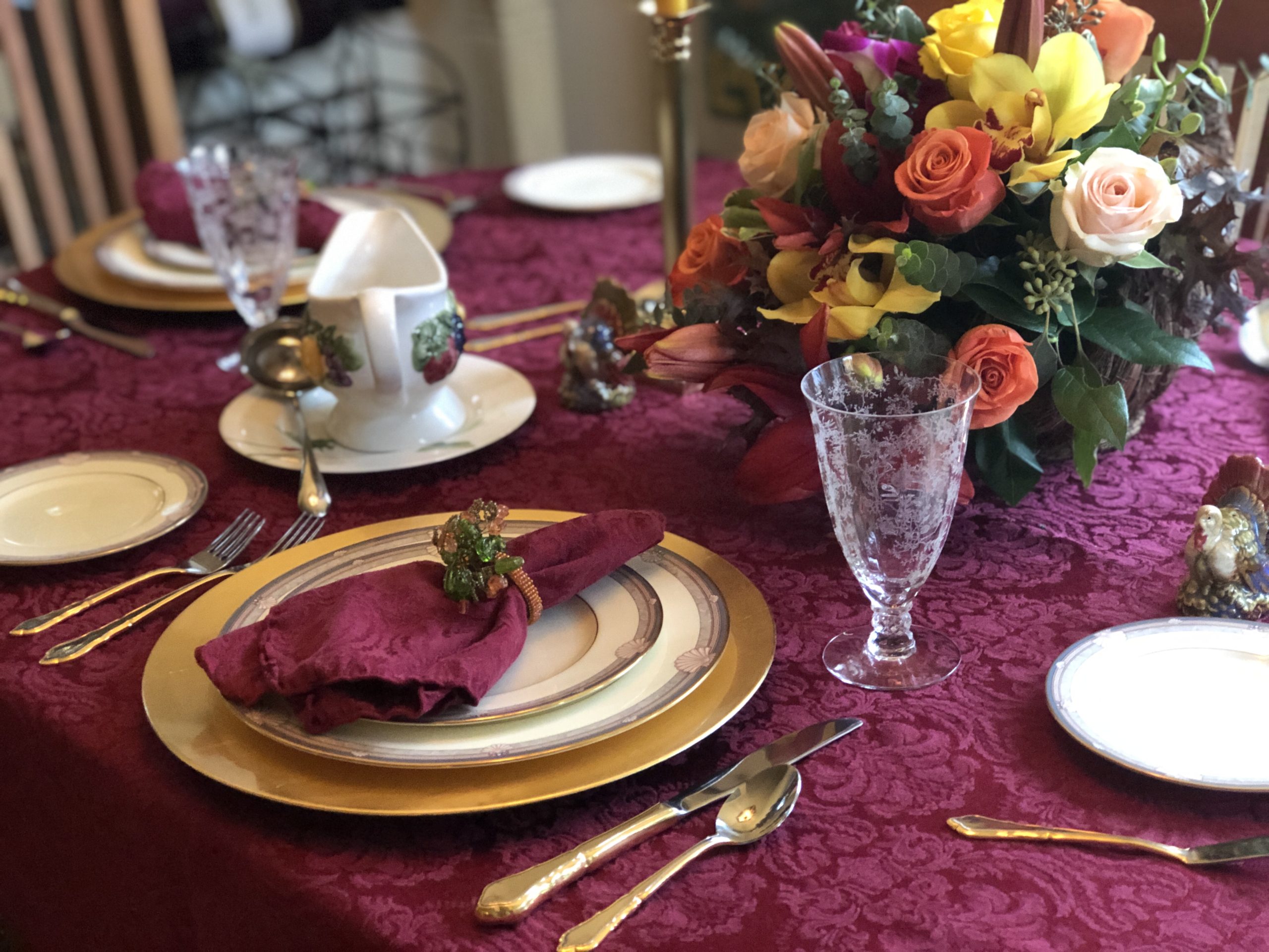 A festive table laid with gold-rimmed plates, a folded burgundy napkin, glassware and a vase of roses