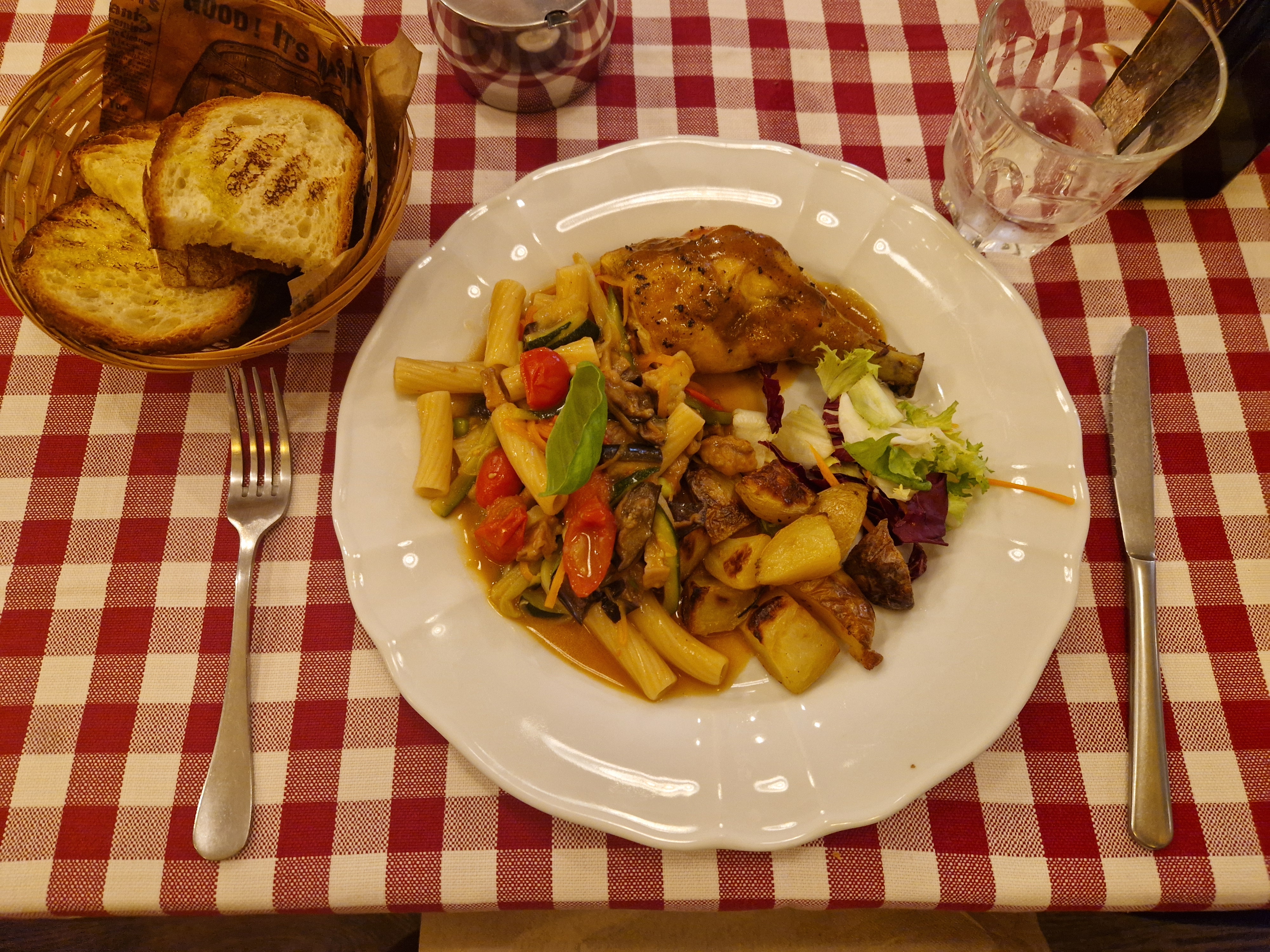 An individual plate of pasta, roast chicken, potatoes and salad on a red checked tablecloth with a bread basket