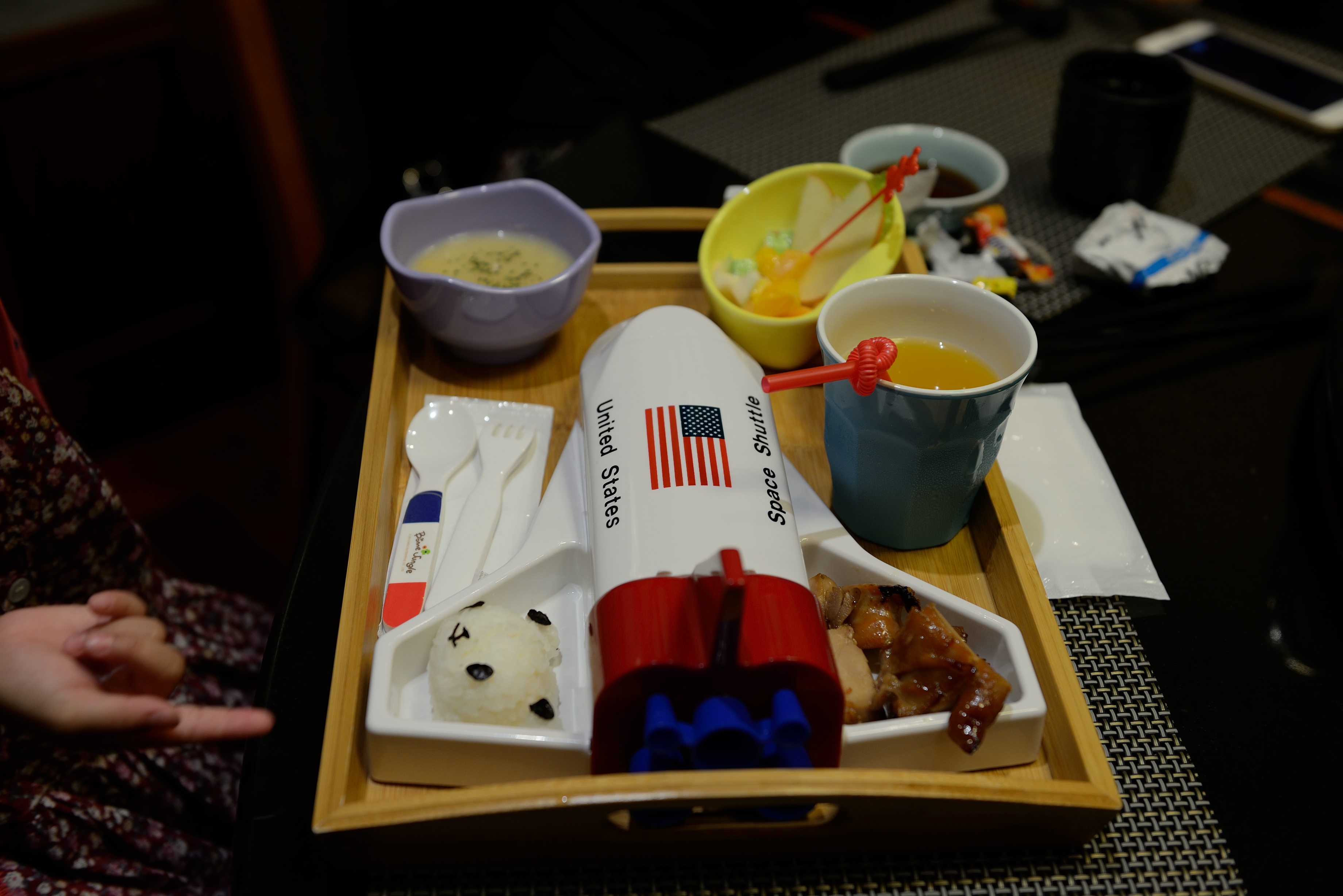 A child's restaurant meal on a wooden tray with soup, fruit, juice and rice