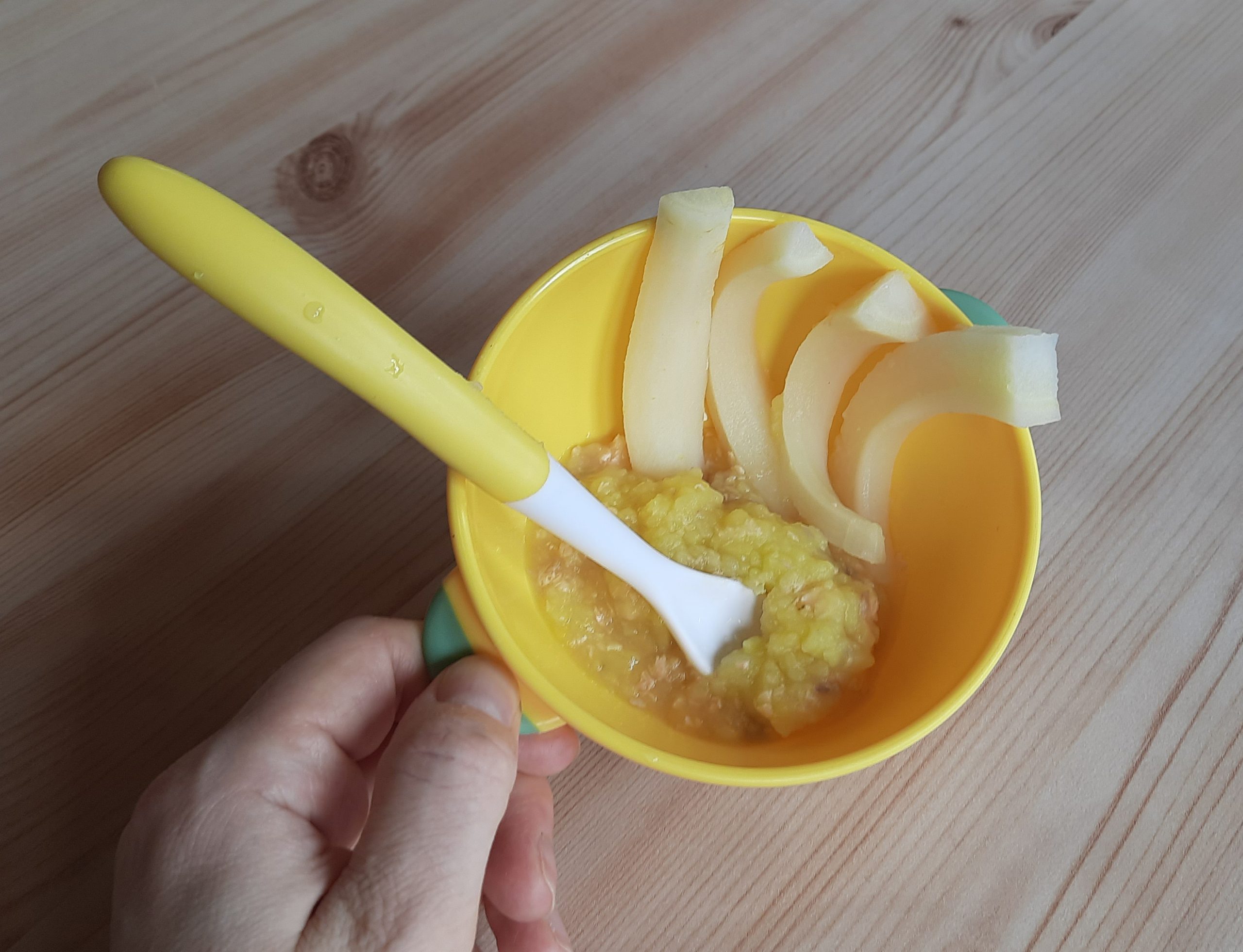 A toddler's yellow bowl and soft-tipped spoon with mashed food and apple slices on a wooden table