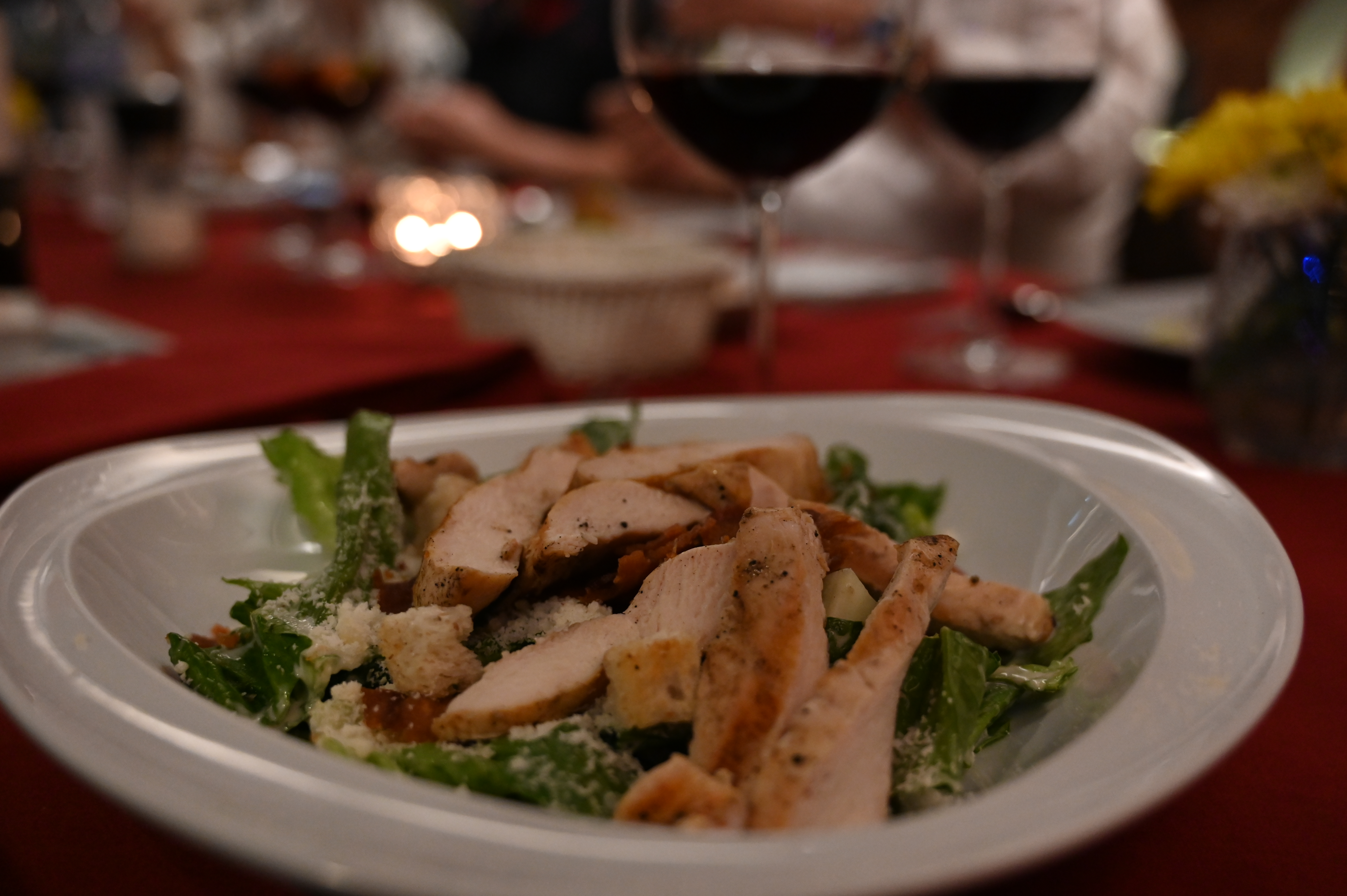 A chicken salad in the foreground of a candlelit dinner table with glasses of red wine behind