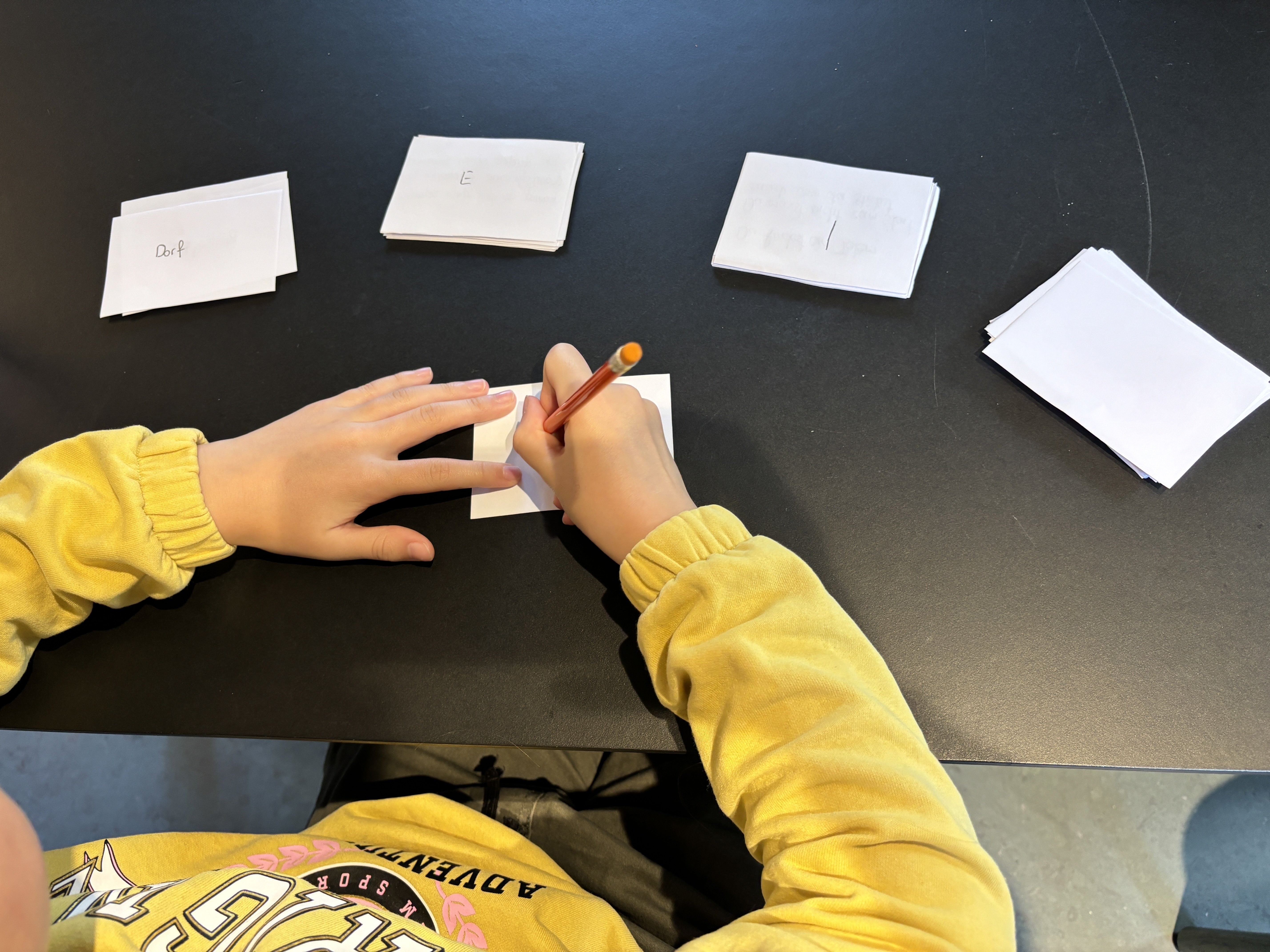A child's hands writing on small paper cards laid out on a dark table for a word game