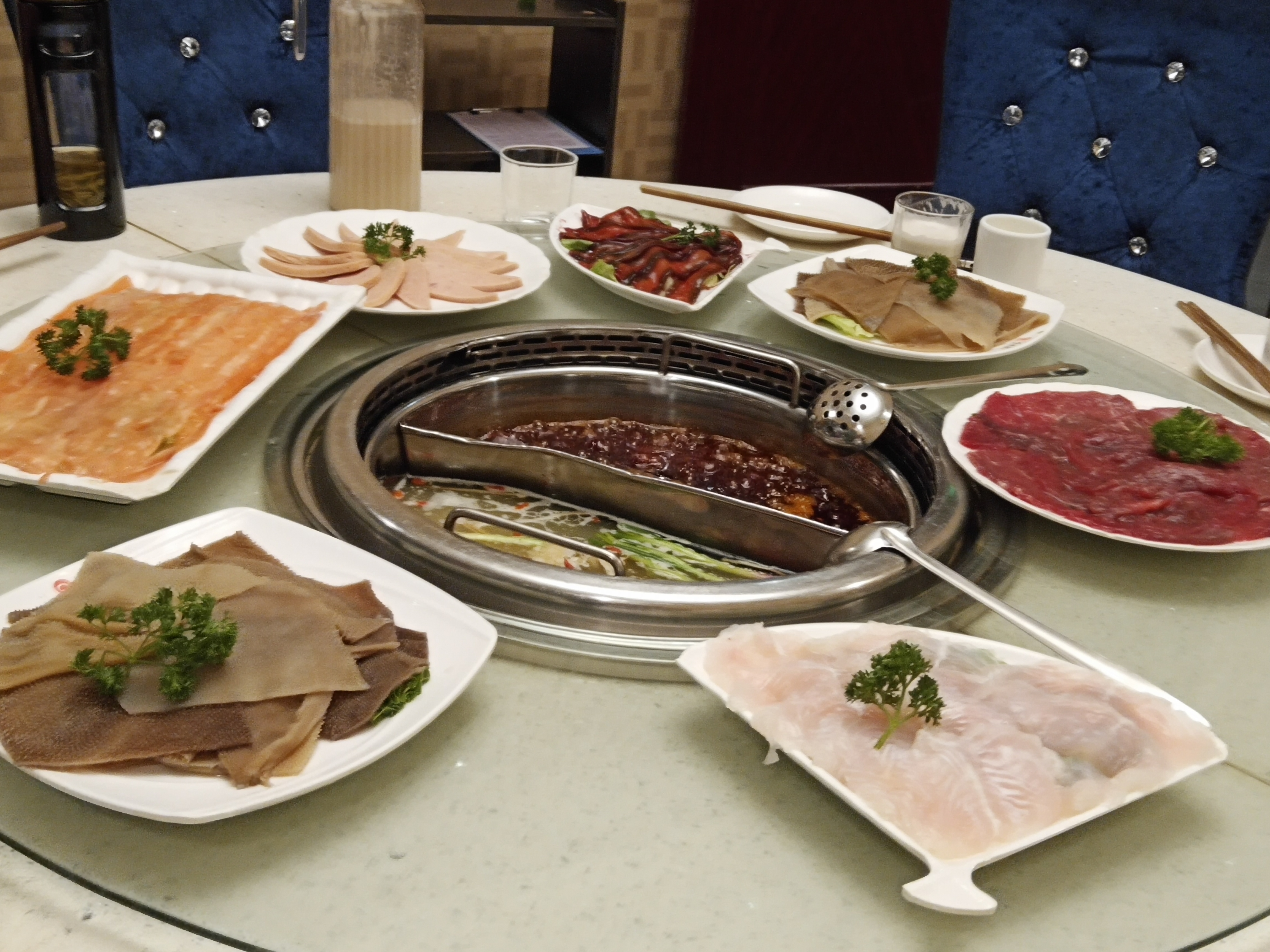 A round table set for a shared hotpot meal with plates of sliced meat, fish and vegetables around a central pot