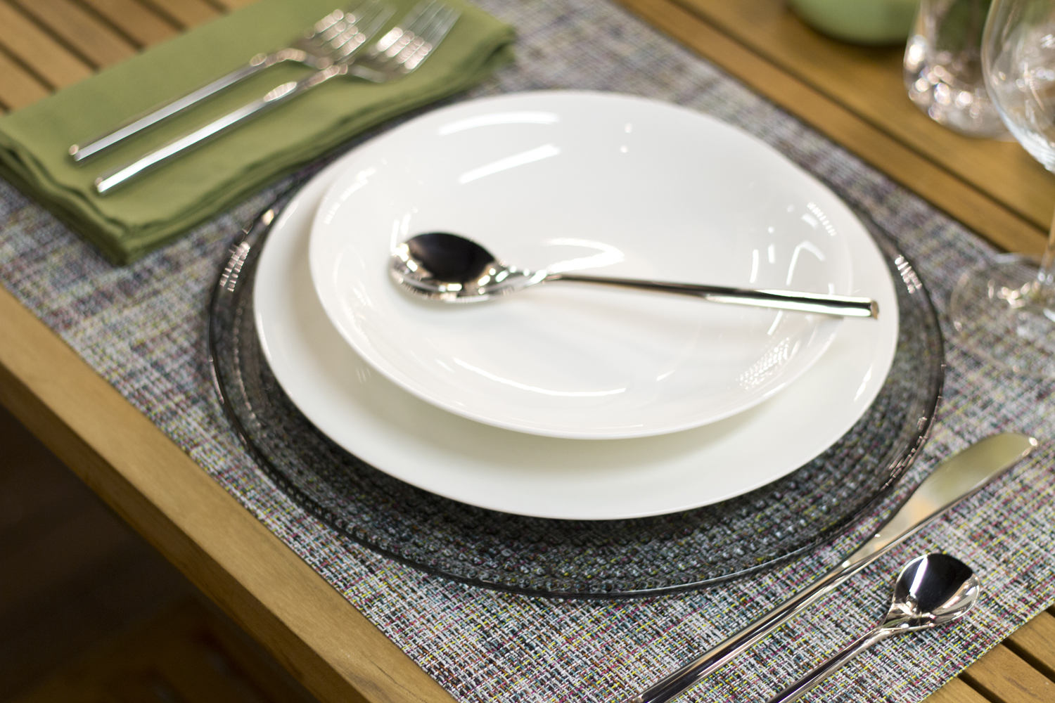 A single place setting with a white soup plate, spoon, cutlery and green napkin on a woven placemat