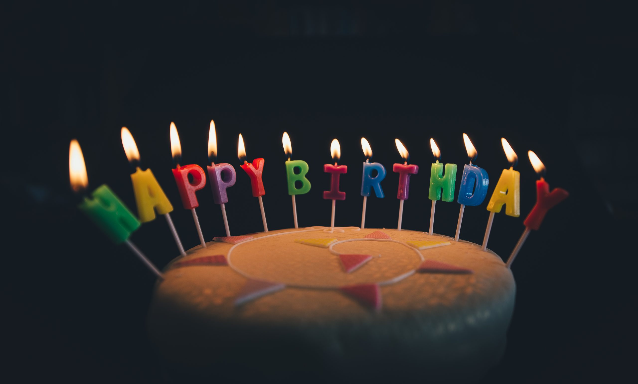 Lit candles spelling Happy Birthday in coloured letters on top of a cake against a dark background