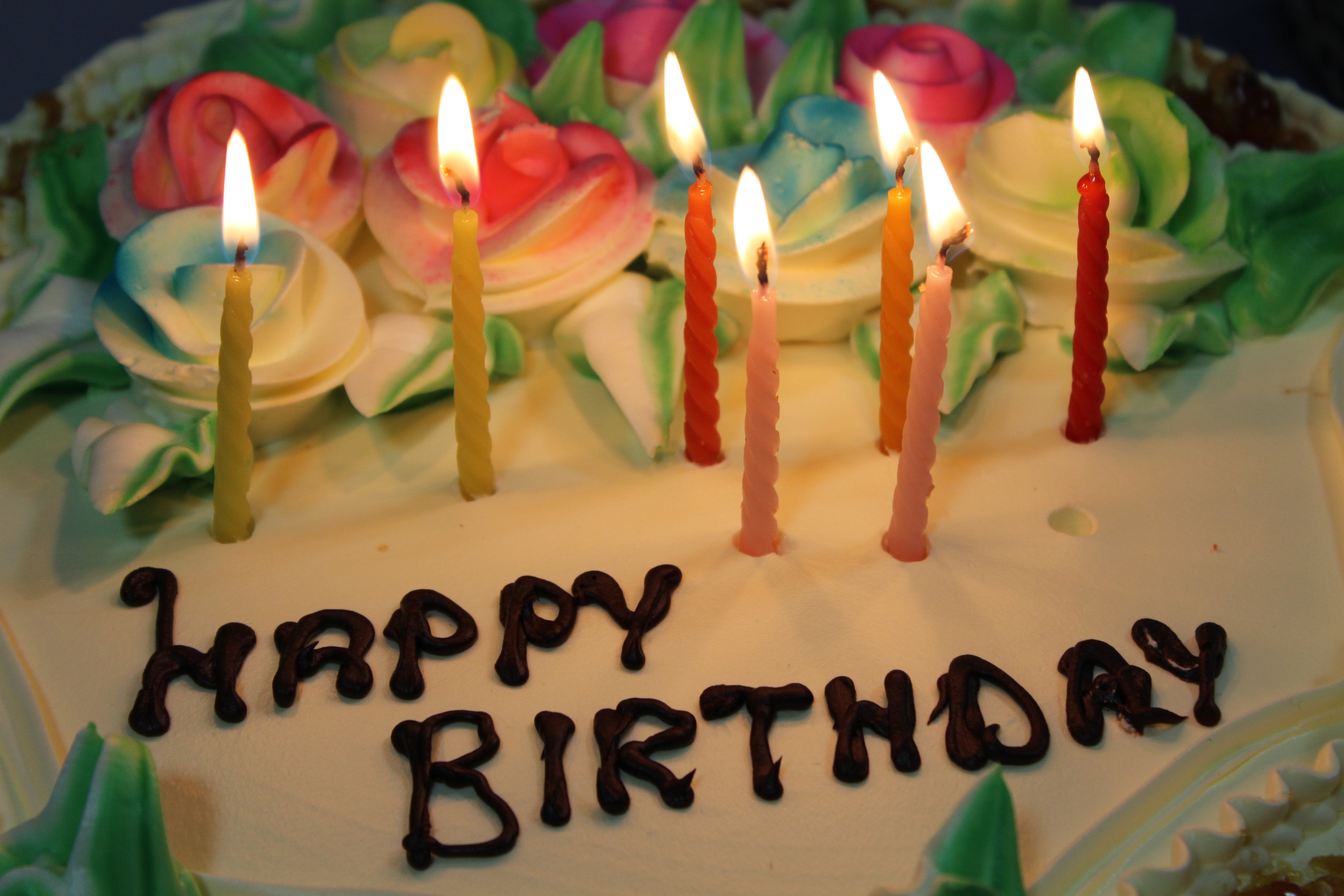 A cream birthday cake decorated with piped roses and seven lit candles above chocolate lettering