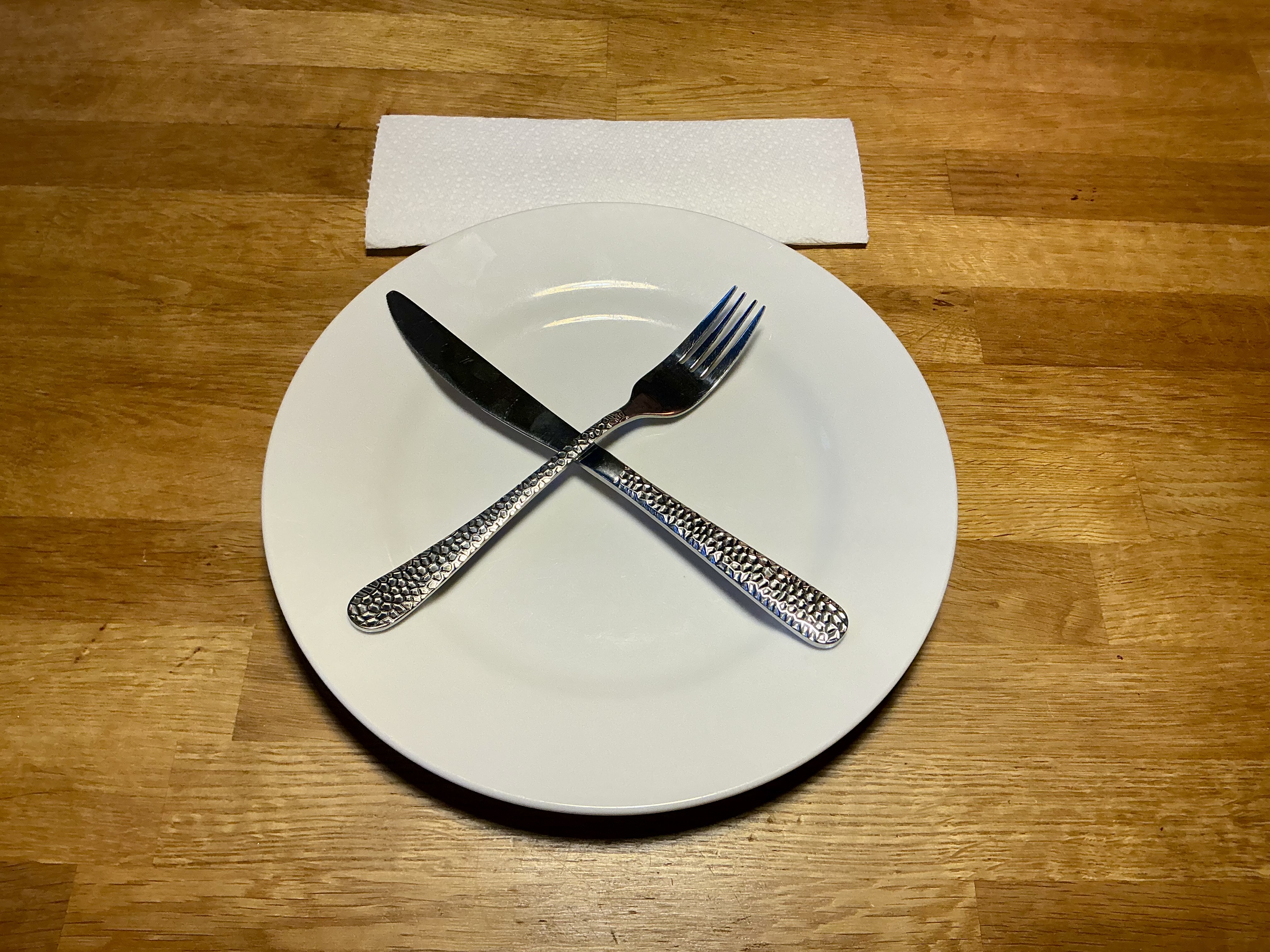 A white plate on a wooden table with a knife and fork crossed in the middle and a paper napkin above