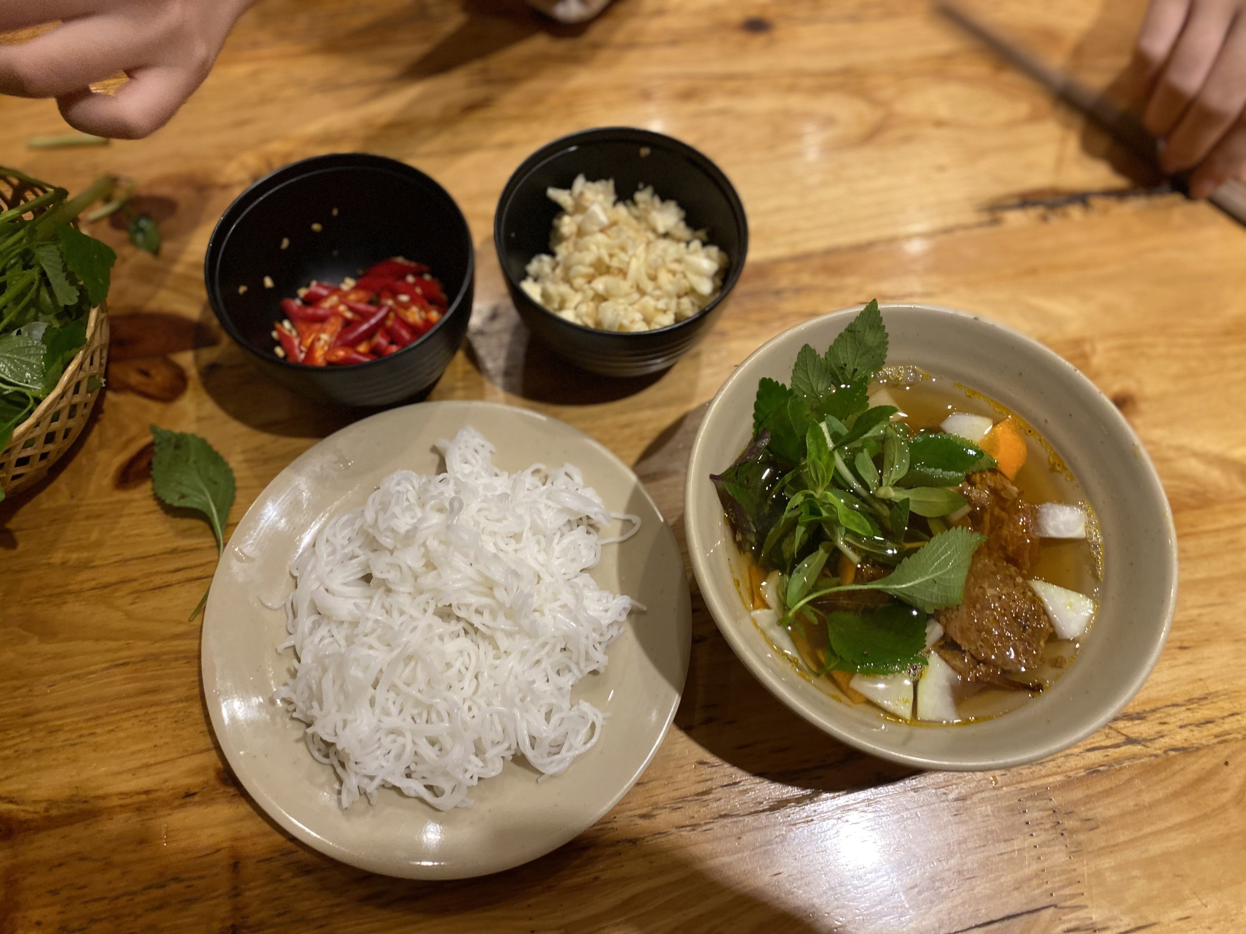 Bowl of noodles, broth with herbs, and side dishes of chopped chilli and garlic on a wooden table