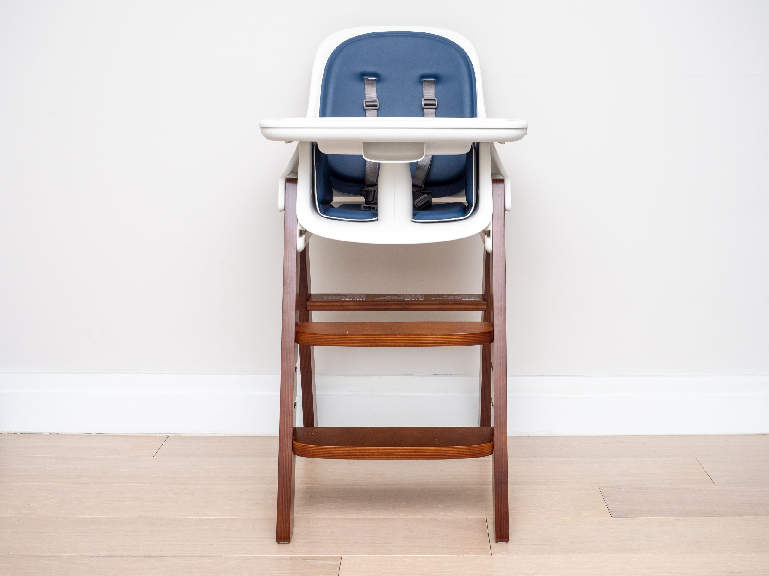 Wooden high chair with a navy seat and white tray standing against a pale wall