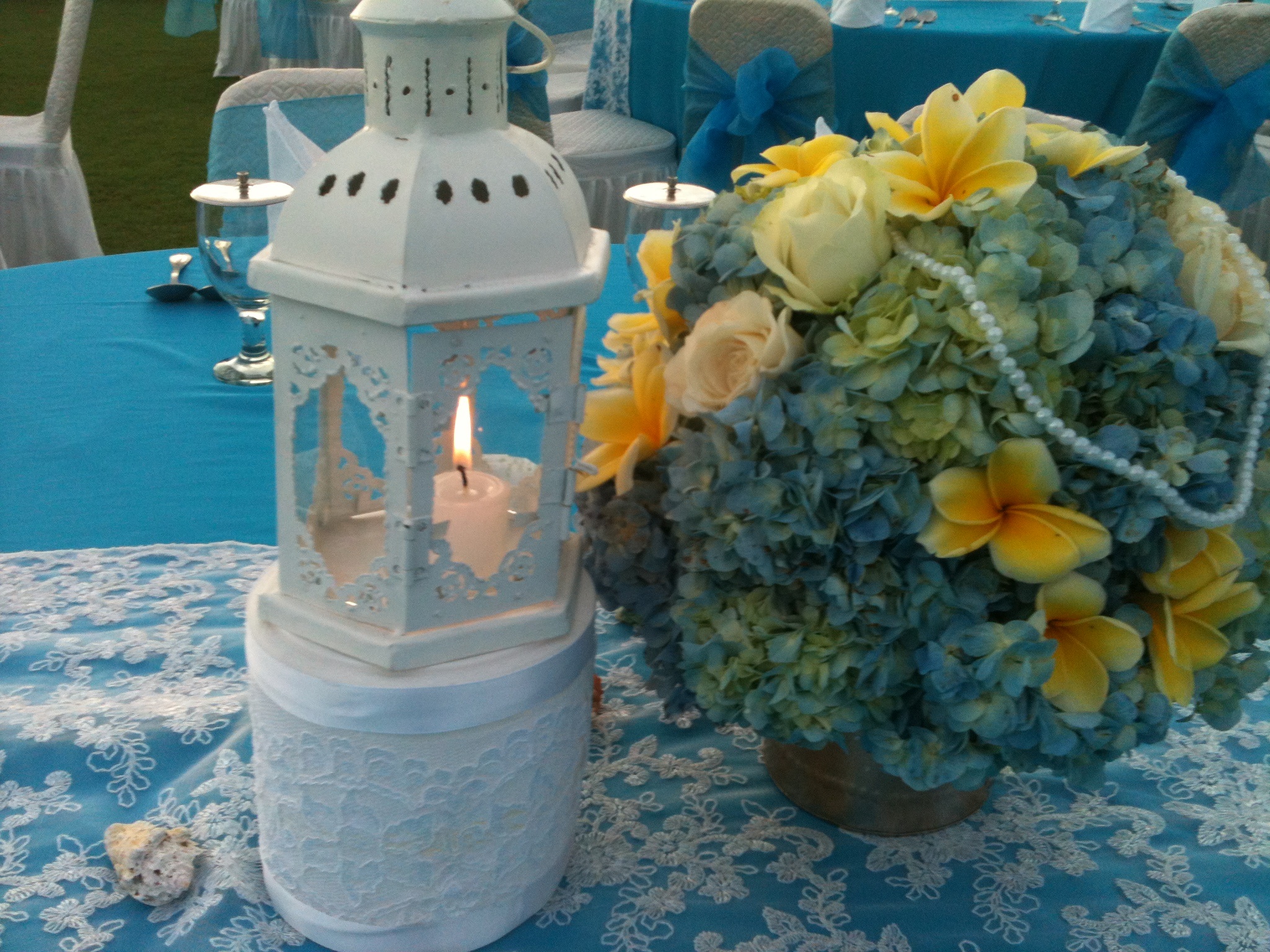 Blue hydrangea and cream rose centrepiece beside a lantern candle on a blue tablecloth