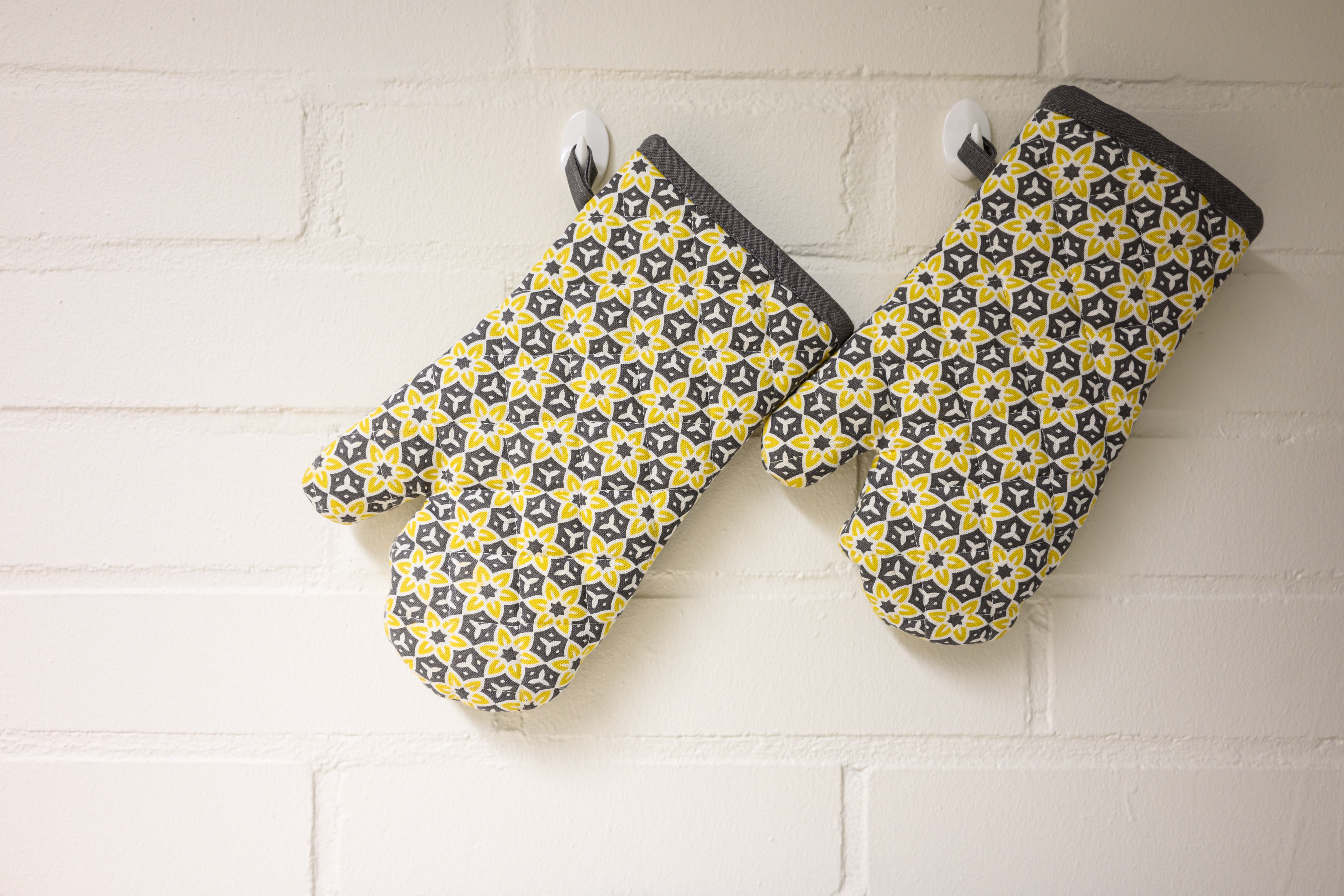Two patterned oven gloves hanging on hooks on a white kitchen wall