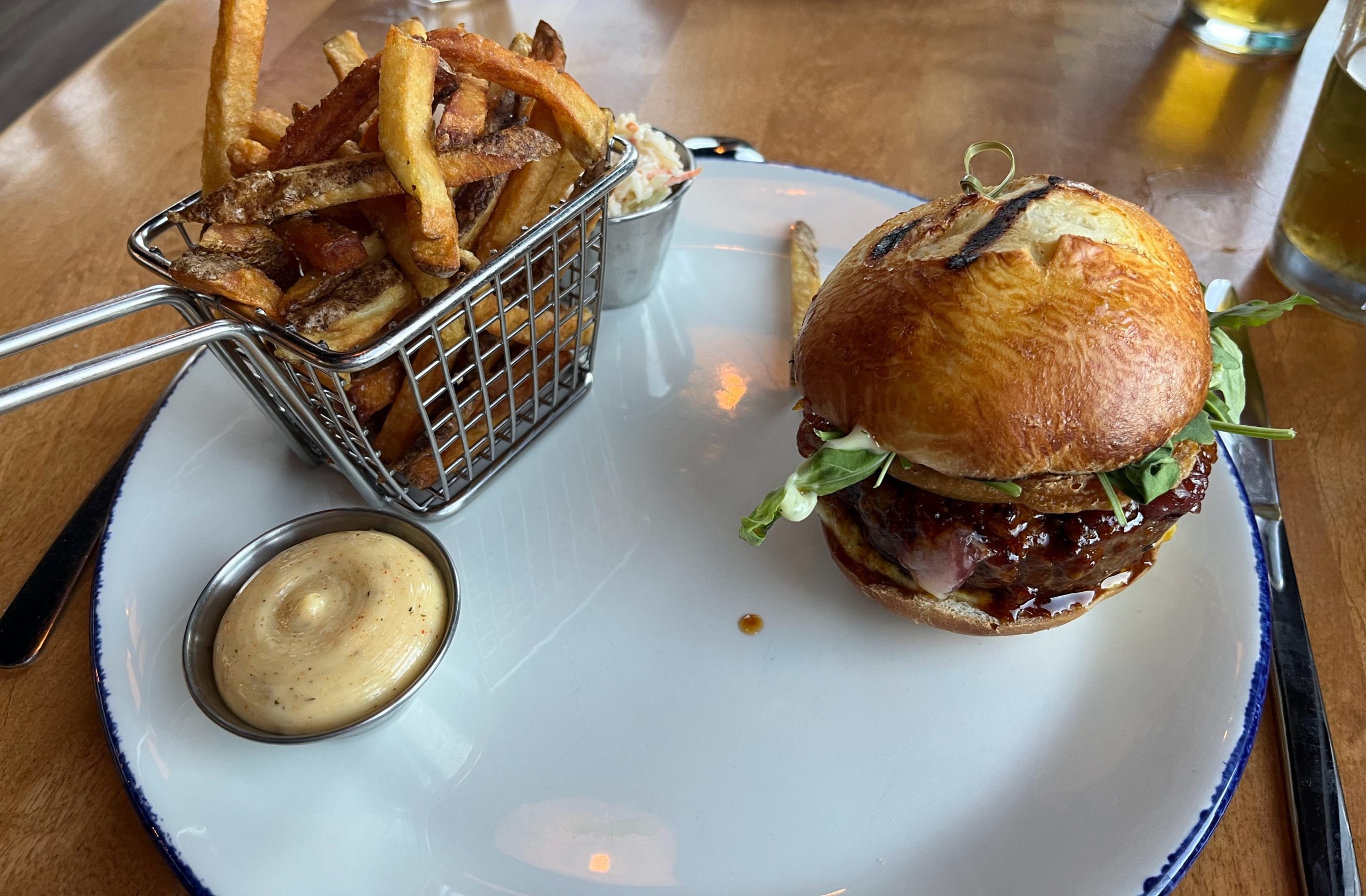 A burger in a brioche bun beside a basket of fries and a pot of aioli on a restaurant table