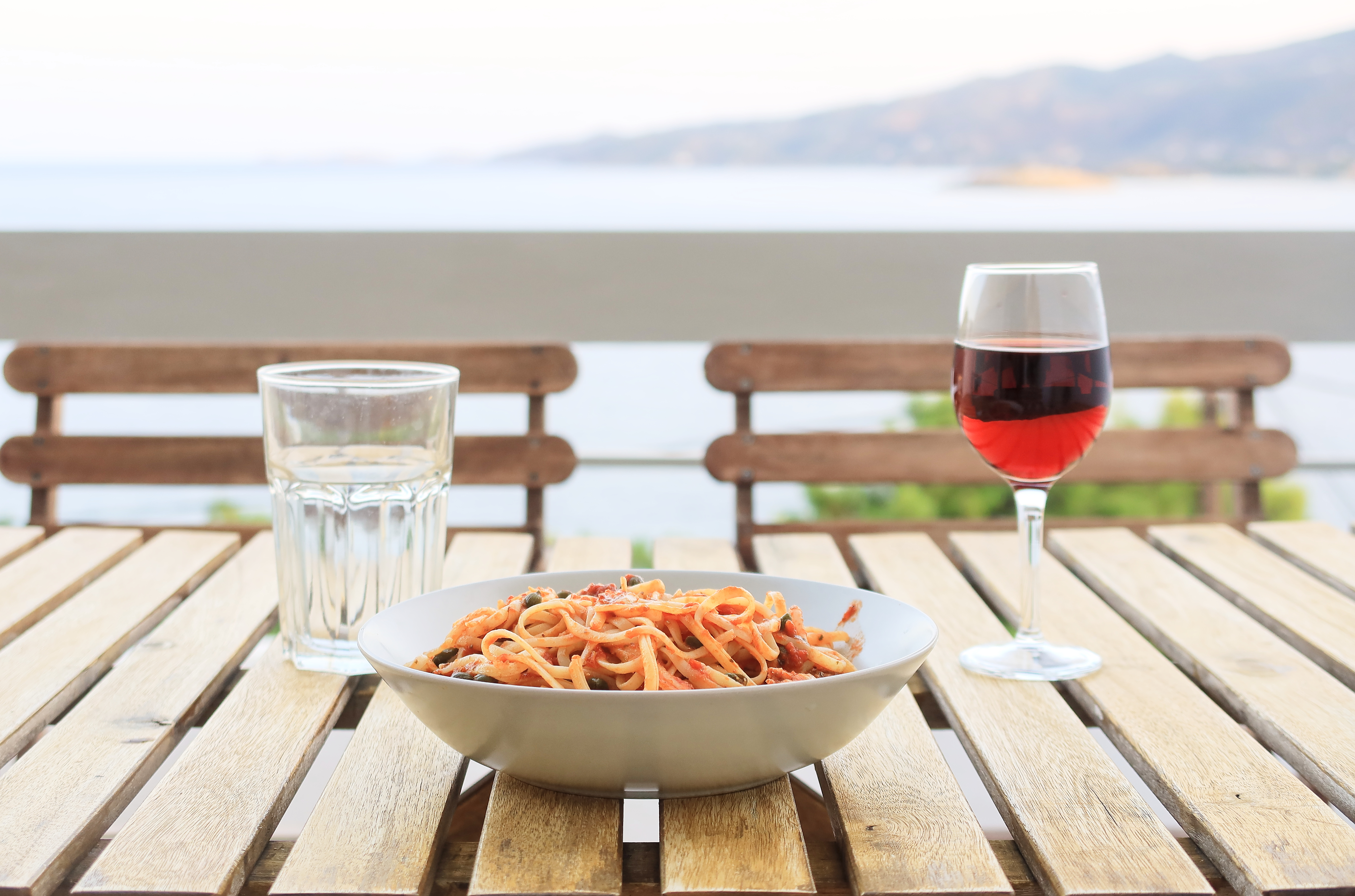 A bowl of pasta with tomato sauce on a wooden table outdoors, with glasses of water and wine