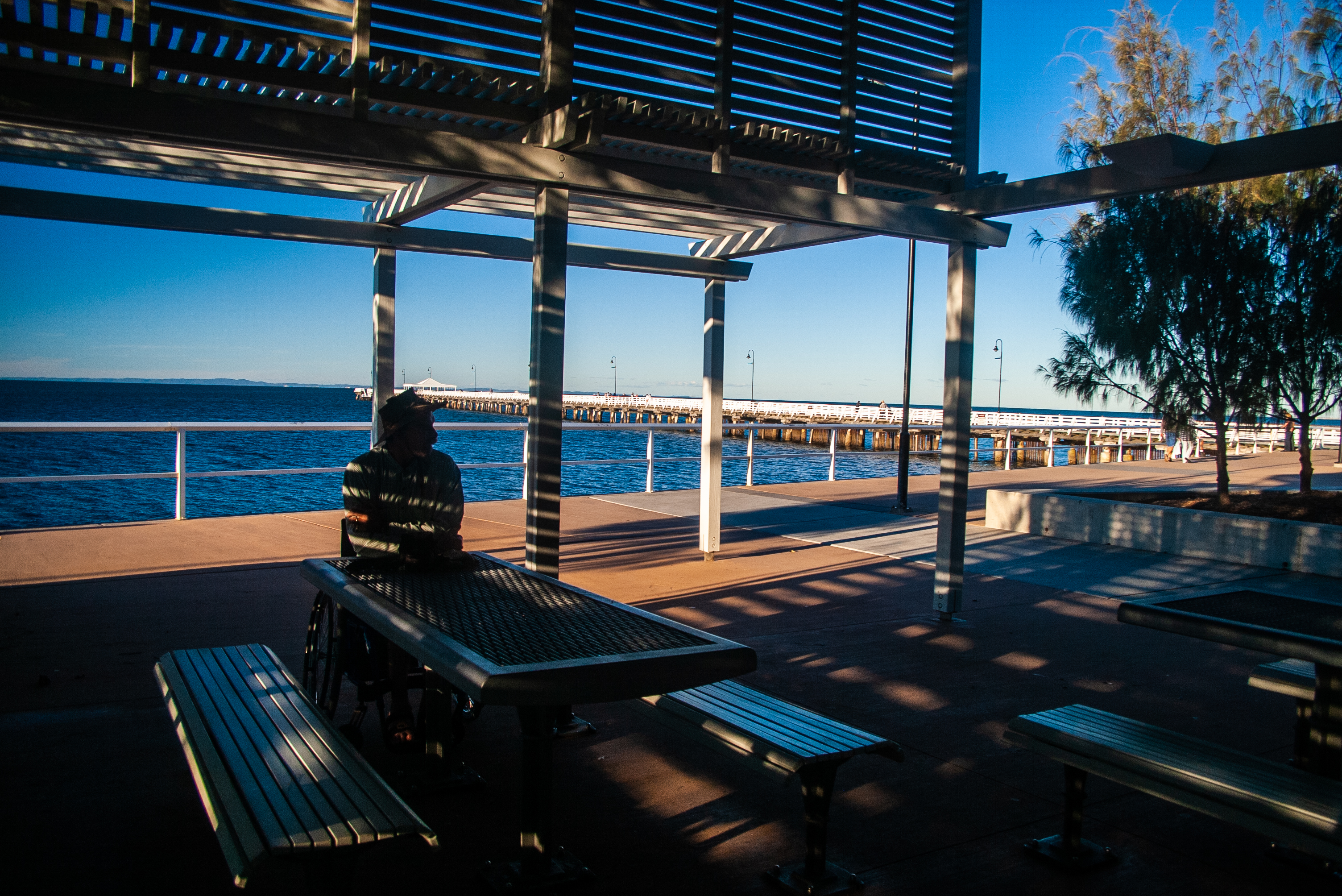 Wheelchair user seated at the extended end of an outdoor table in a shaded seafront shelter