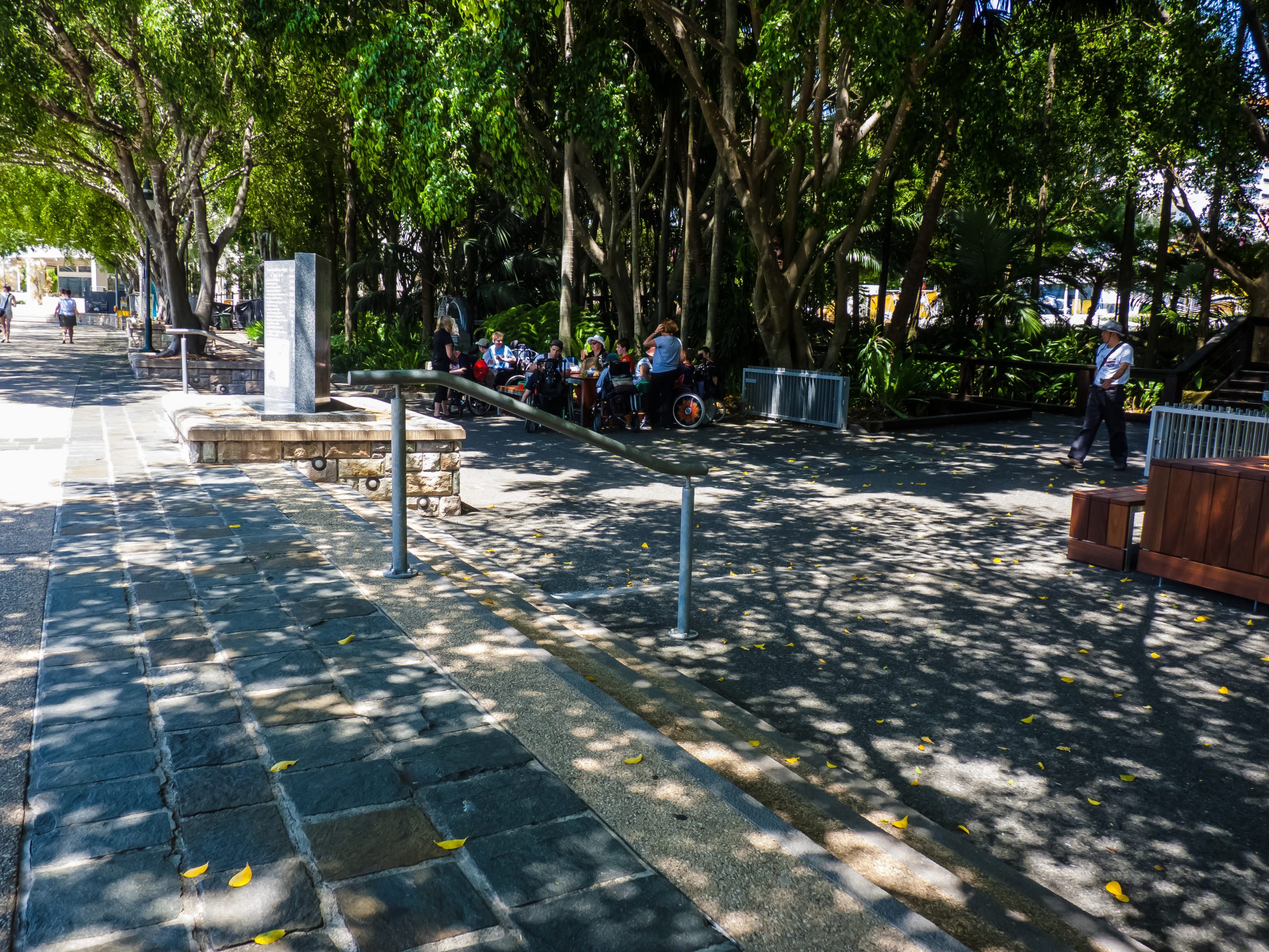 A wide, level paved path with a handrail leading to shaded outdoor tables where wheelchair users are gathered